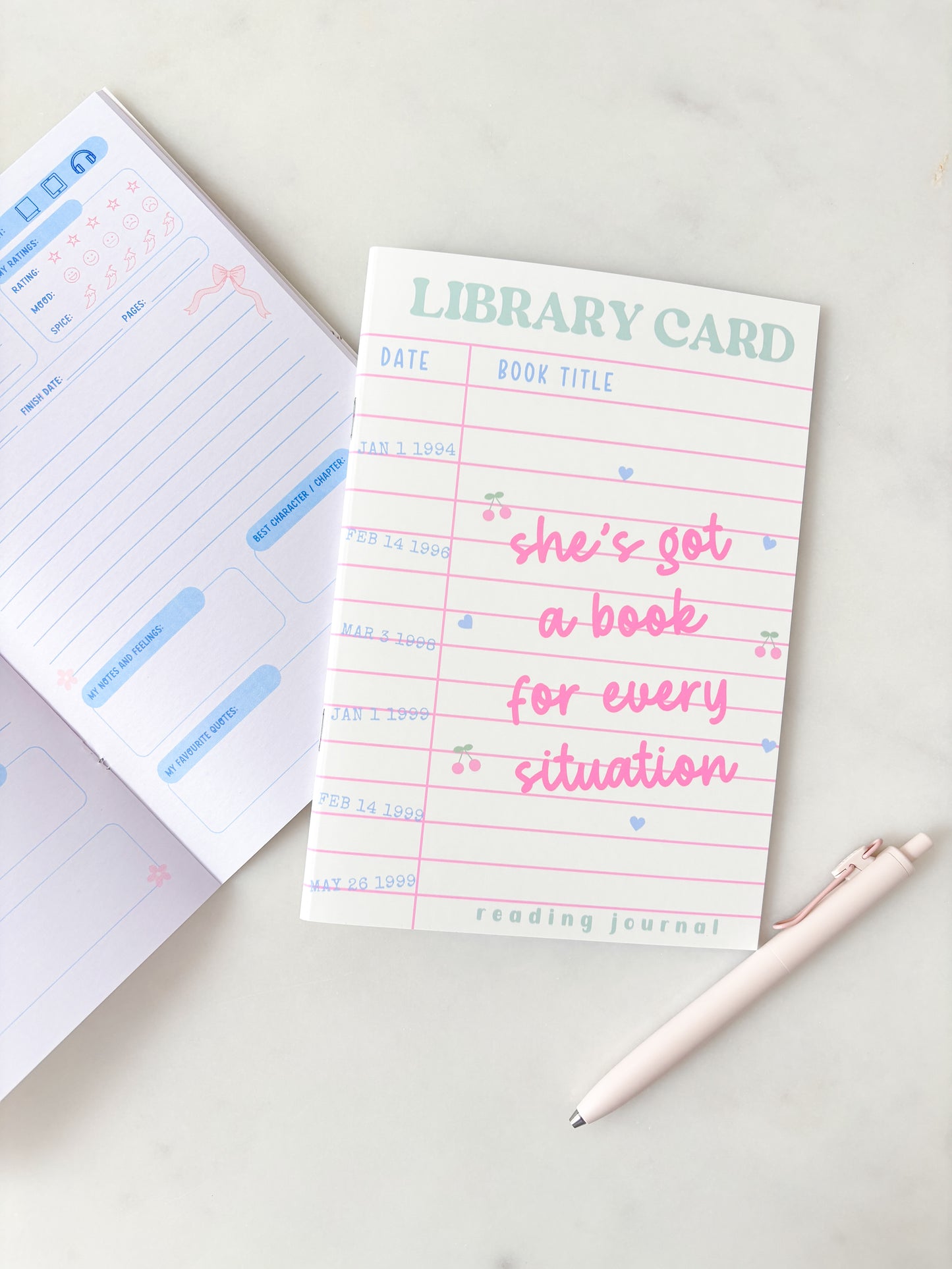 A reading journal notepad titled 'LIBRARY CARD' with lined pages, pink and blue text, and a cute phrase, styled on a light marble surface with an open planner and a pen.