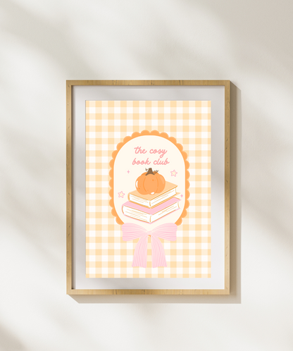 A framed art print featuring a cute illustration of a pumpkin on a stack of books with the text 'the cozy book club' on a yellow-and-white gingham background, displayed on a light wall with soft shadow overlays.